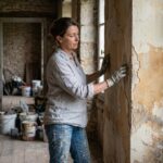 Femme inspectant une peinture ancienne qui s'écaille sur un mur en plâtre dans une vieille maison avant travaux de rénovation